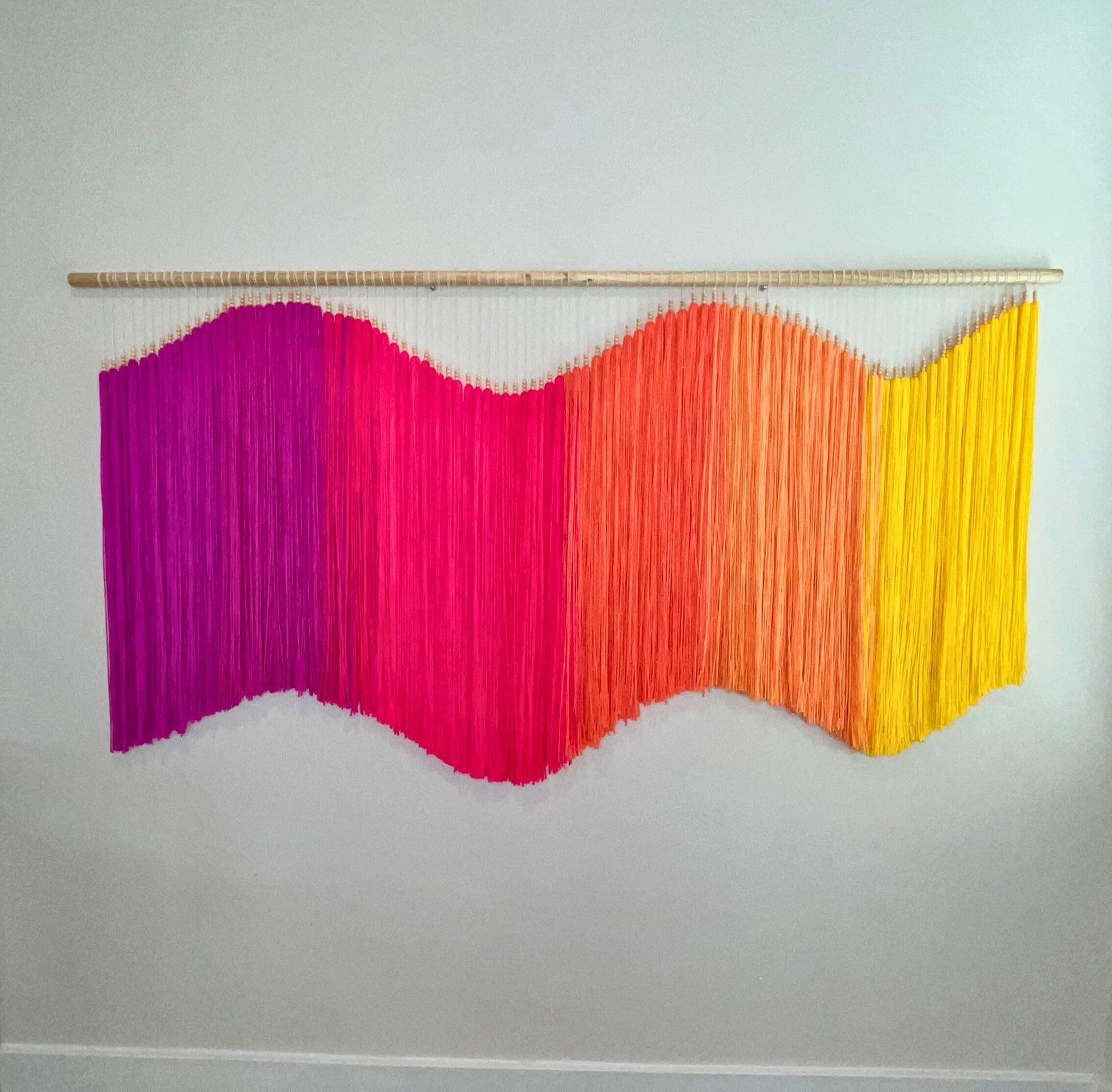 Colorful yarn fiber art macramé wall hanging with fuchsia, pink, orange, and yellow strands on a white wall. Statement piece, focal art, focal piece, sensory art, dimensional art, kinetic art, textural art, textured art. 