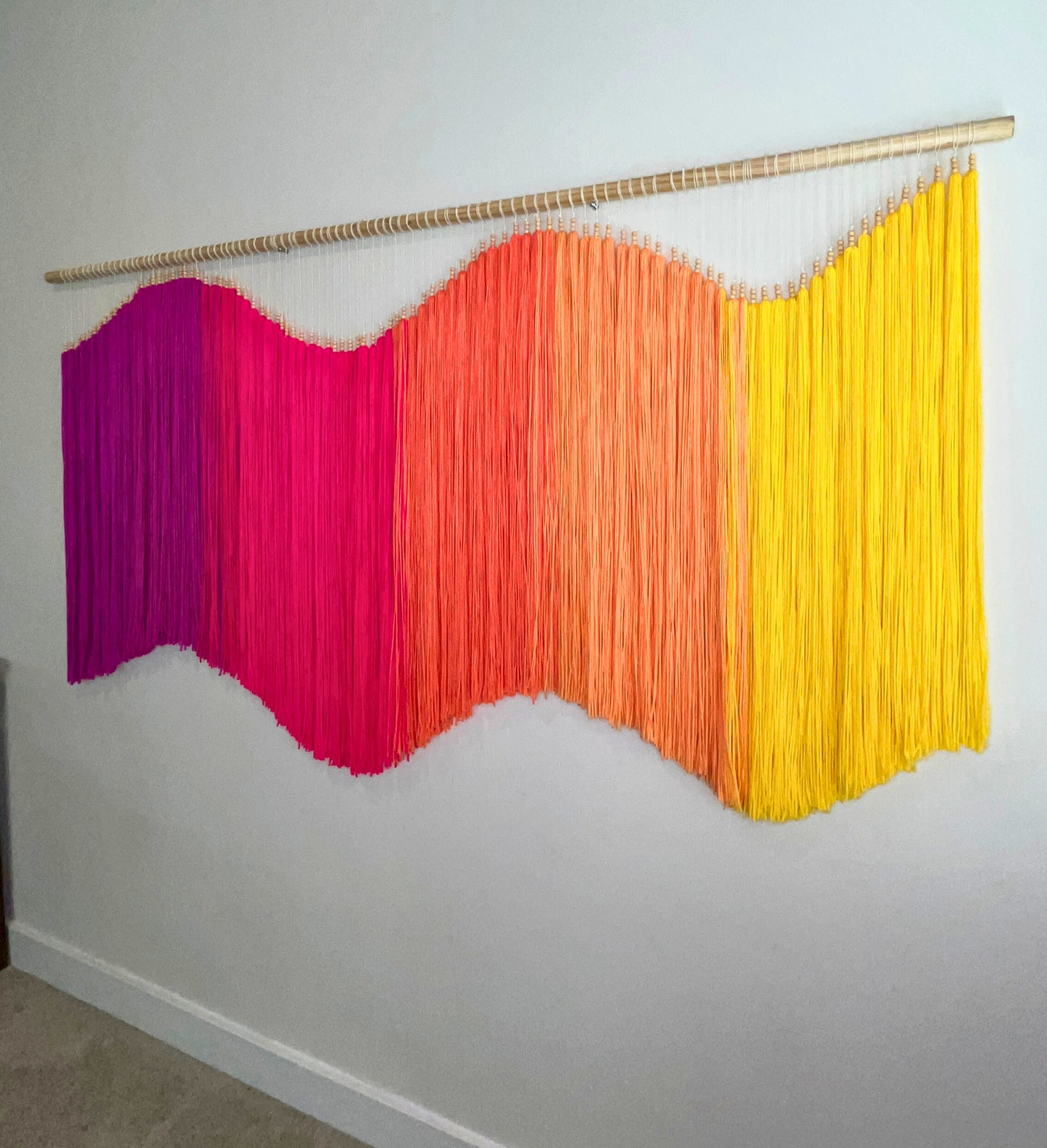 Colorful yarn fiber art macramé wall hanging with fuchsia, pink, orange, and yellow strands on a white wall. Statement piece, focal art, focal piece, sensory art, dimensional art, kinetic art, textural art, textured art. 