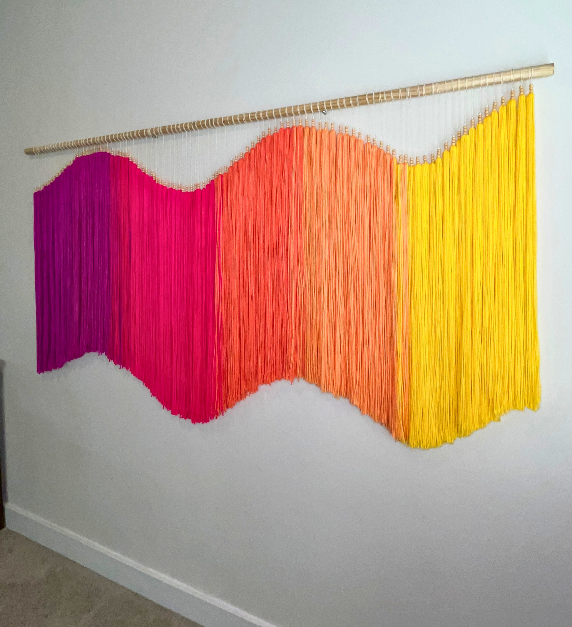 Colorful yarn fiber art macramé wall hanging with fuchsia, pink, orange, and yellow strands on a white wall. Statement piece, focal art, focal piece, sensory art, dimensional art, kinetic art, textural art, textured art. 