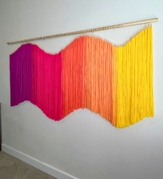 Colorful yarn fiber art macramé wall hanging with fuchsia, pink, orange, and yellow strands on a white wall. Statement piece, focal art, focal piece, sensory art, dimensional art, kinetic art, textural art, textured art. 