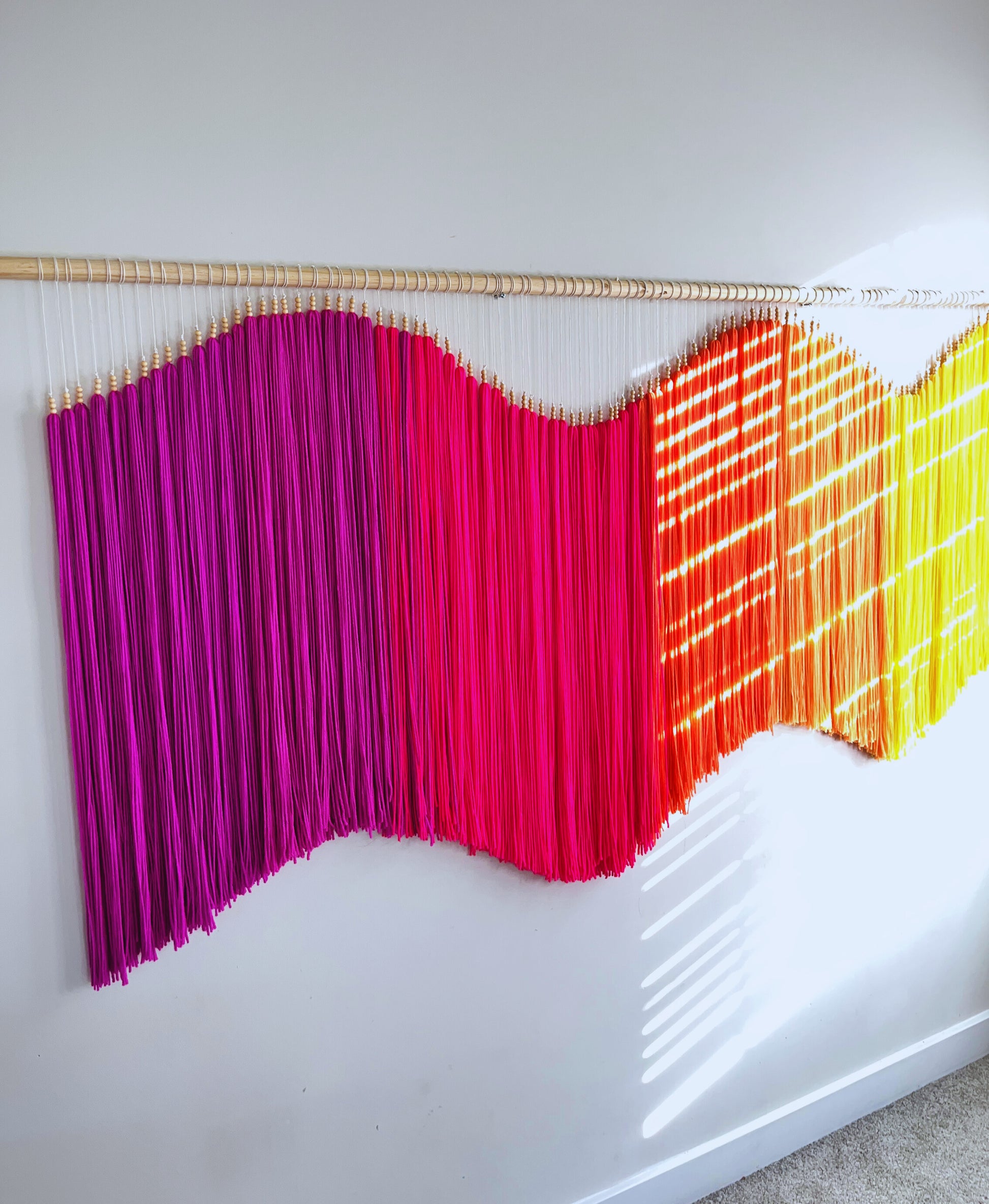 Colorful yarn fiber art macramé wall hanging with fuchsia, pink, orange, and yellow strands on a white wall. Statement piece, focal art, focal piece, sensory art, dimensional art, kinetic art, textural art, textured art. 